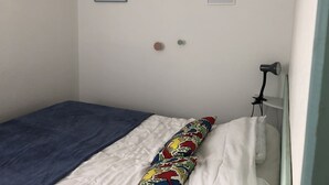 Un petit lit bien fait, avec une couverture bleue et un coussin coloré, un mur blanc orné de deux tableaux encadrés et une petite lampe posée sur une table de chevet.
