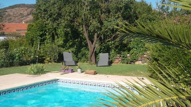 Una piscina con dos sillas de descanso, rodeada de exuberante vegetación y un muro de piedra.