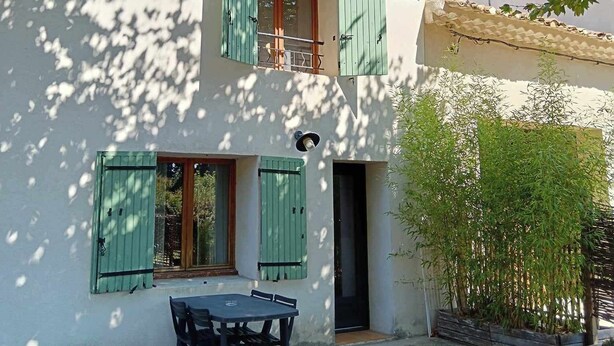 Un edificio blanco con contraventanas verdes, un balcón y una mesa con sillas afuera.