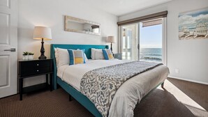 A bedroom with a bed, a nightstand, a lamp, a window with a view of the sea, and a painting on the wall.