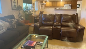 Ein Wohnzimmer mit braunem Ledersofa, einem Glastisch mit Büchern und einer Fernbedienung, und einer Küche im Hintergrund.