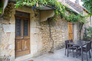 Un edificio de piedra con puerta y ventana de madera, una mesa con sillas y un muro cubierto de hiedra.