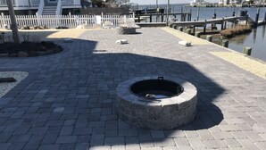 A paved waterfront area with a circular stone feature, a white picket fence, and a dock extending into the water. - Bayfront with dock and fire pit, @ Harris Harbor, Beach  Haven Park, LBI (Long Beach Township)