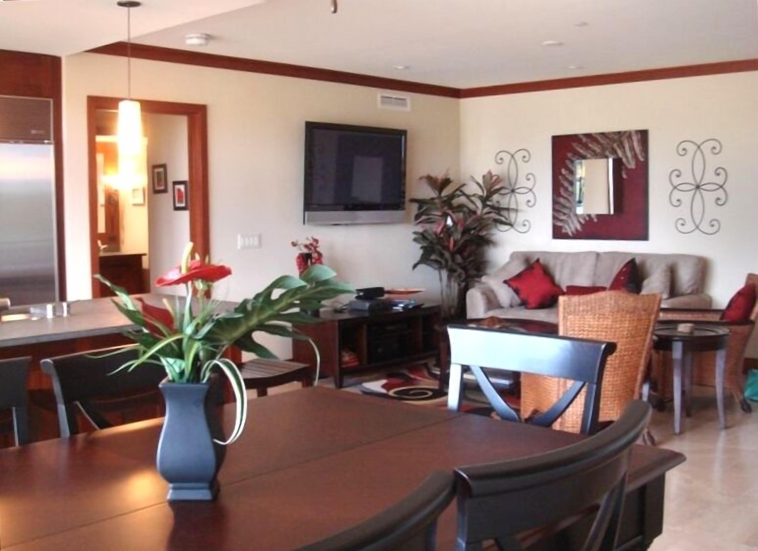 A modern living room with a dining area, a flat-screen TV mounted on the wall, and a large potted plant.