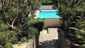 Un chemin de pierres mène à une piscine entourée d’épais arbres à feuilles persistantes.