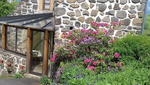 Un bâtiment en pierre doté d’une entrée en verre, entouré d’une grande variété de fleurs et de plantes.
