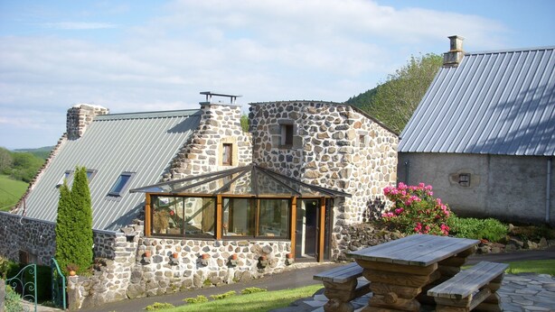 Une maison en pierre avec un toit en pente, une véranda en verre et une terrasse en pierre avec une table en bois.