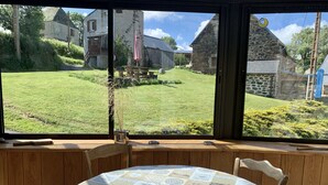 Une pièce dotée d’une grande fenêtre donnant sur une cour verdoyante et des bâtiments en pierre. Une table ronde, recouverte d’une nappe à motifs, est placée près de la fenêtre.