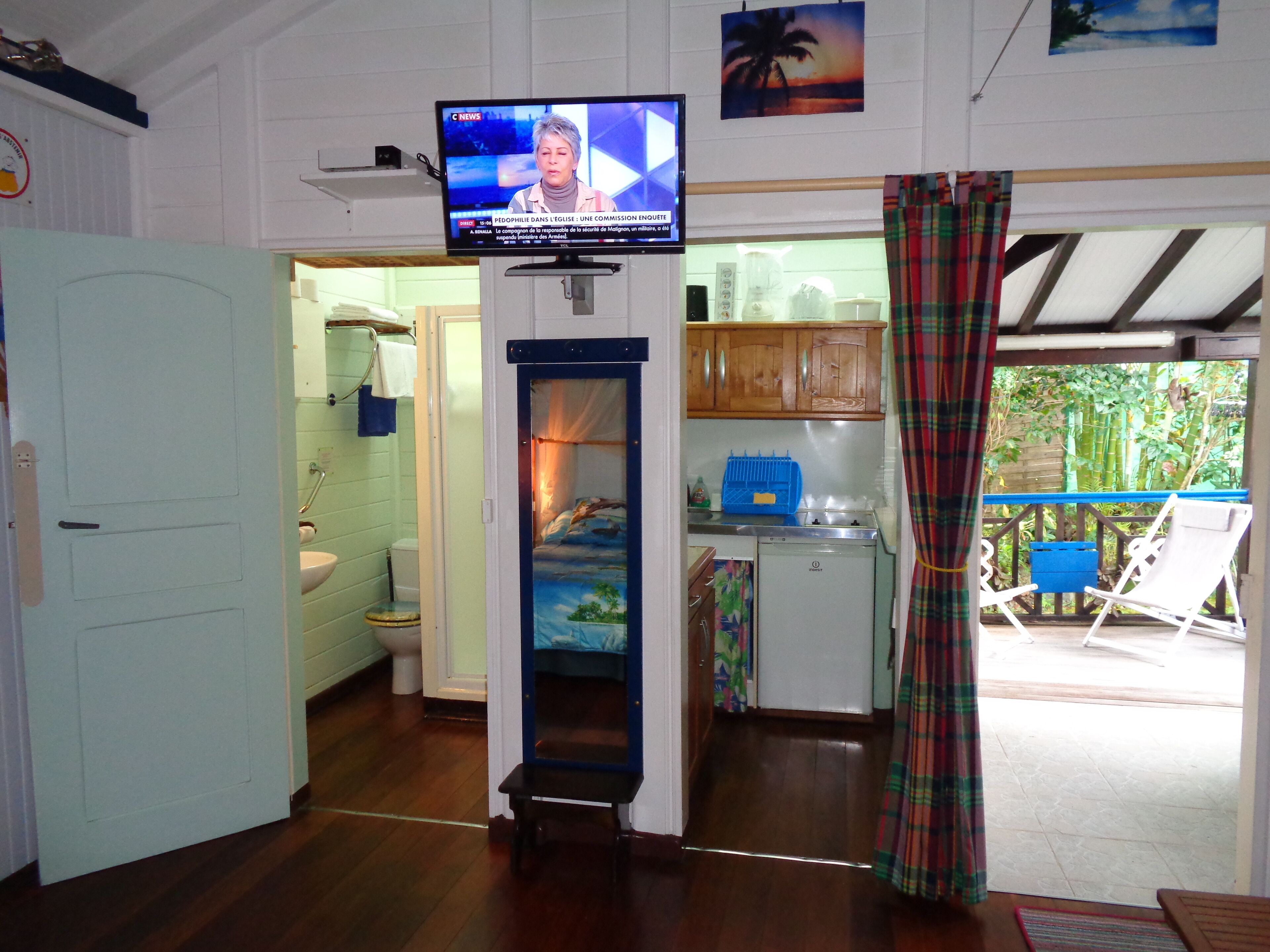 Un espace de vie compact doté d’un téléviseur fixé au mur, d’une petite kitchenette et d’une vue sur un espace extérieur accessible par une porte ouverte.