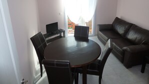 En stue med en brun lædersofa, et rundt spisebord med fire stole, et fladskærms-tv på et stativ og en skydedør til et udendørs område.