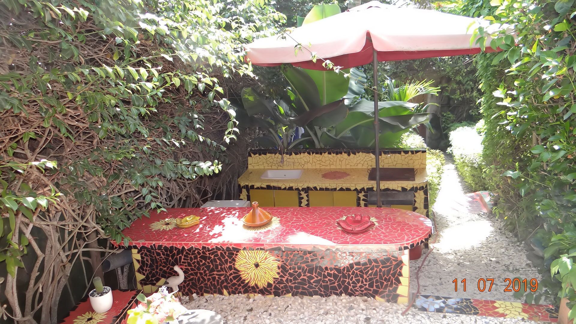 An outdoor dining area with a mosaic-tiled table, a red umbrella, and a stone sink.