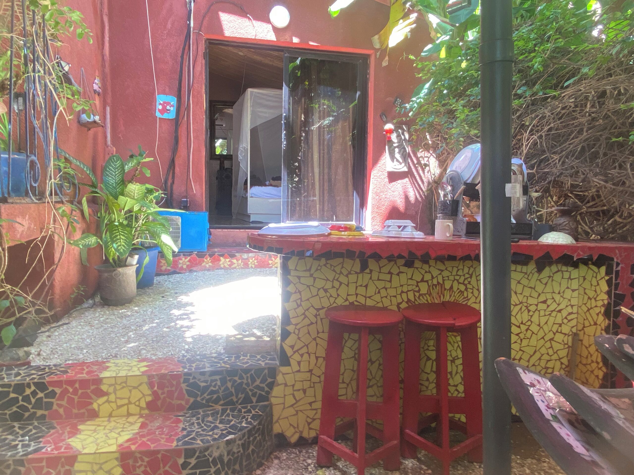 Uma área de pátio com chão em mosaico, um pequeno balcão de bar com banquetas e plantas em vasos.