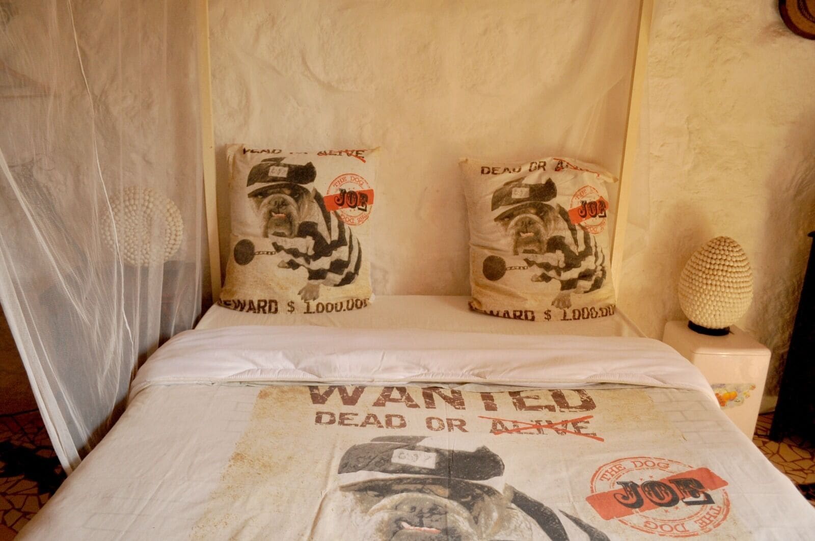 A bed with a 'Wanted Dead or Alive' poster, two pillows with a dog and 'Joe' design, and a textured wall.