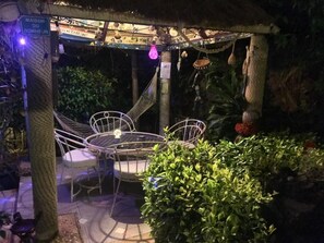Un espacio de descanso al aire libre con techo, que incluye una mesa y sillas metálicas, rodeado de vegetación y una hamaca.