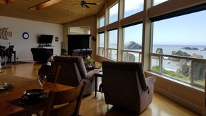 Un salón amplio con techo de madera, ventanas grandes con vistas al mar, zona de comedor con mesa y sillas, y una chimenea.