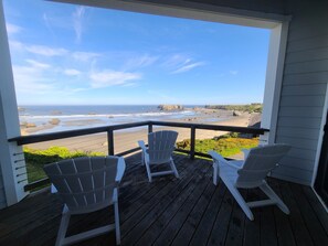 A wooden deck with three white Adirondack chairs facing a beach with rocky outcrops and clear skies.