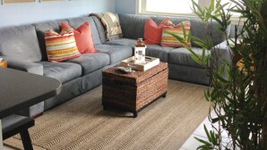 A living room with a grey sofa, a woven coffee table, a potted plant, and a window with blinds. - Beautiful 2nd floor unit with ocean views and 2 minute walk to the beach. (Wrightsville Beach)