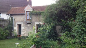 A rustic stone building with a small wooden door and window, surrounded by lush greenery and a flower bed with vibrant flowers. - Charming Cottage Set In The Loire Valley Just Minutes From Sancerre (Feux)