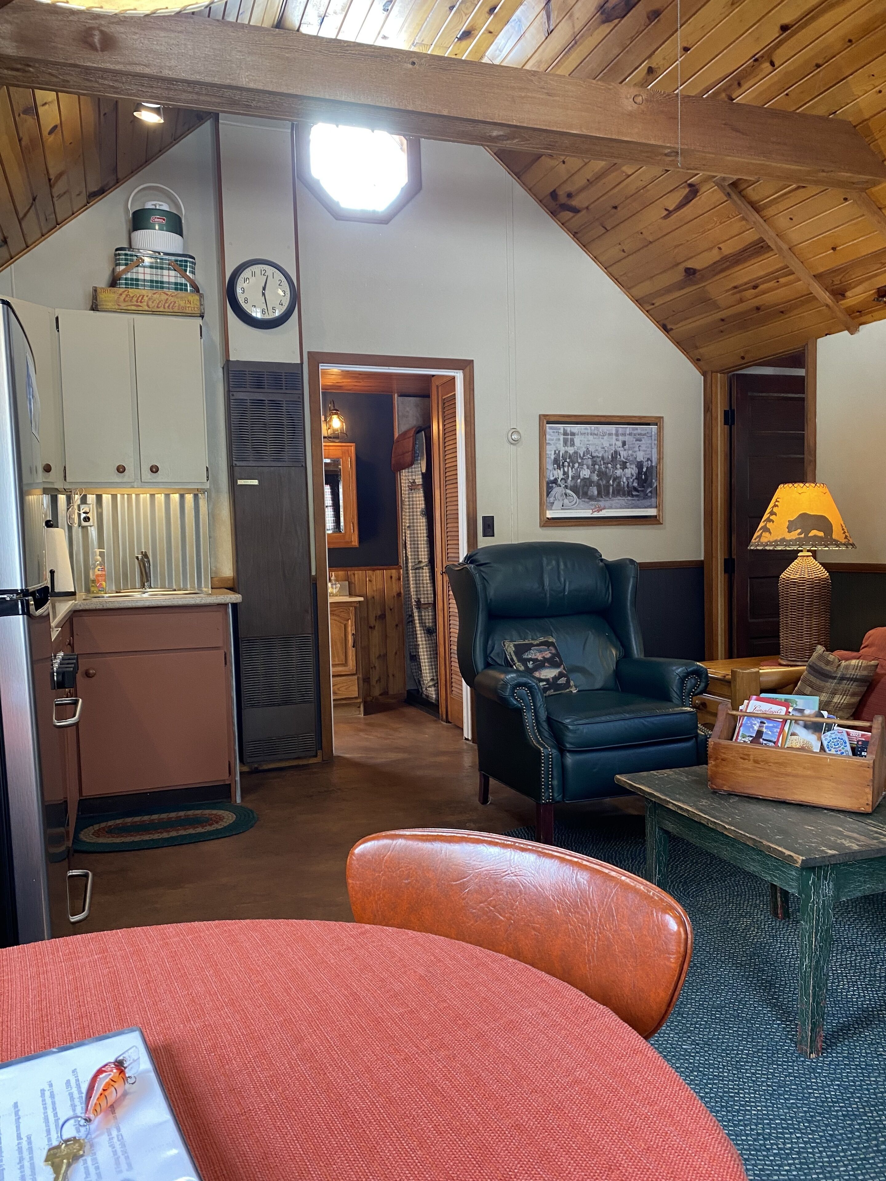A cozy room with a wooden ceiling, a dining table with a red tablecloth, a black leather chair, and a small wooden table with a lamp.
