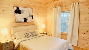 Acogedor dormitorio de cabaña de madera con cama, mesitas de noche, lámpara, un cuadro y ventana con cortinas.