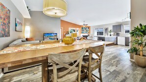 A spacious living area with a wooden dining table, chairs, and a large pendant light. A kitchen with modern appliances and a large window is visible in the background. - Fabulous Location @ Rancho Relaxo  (La Quinta)