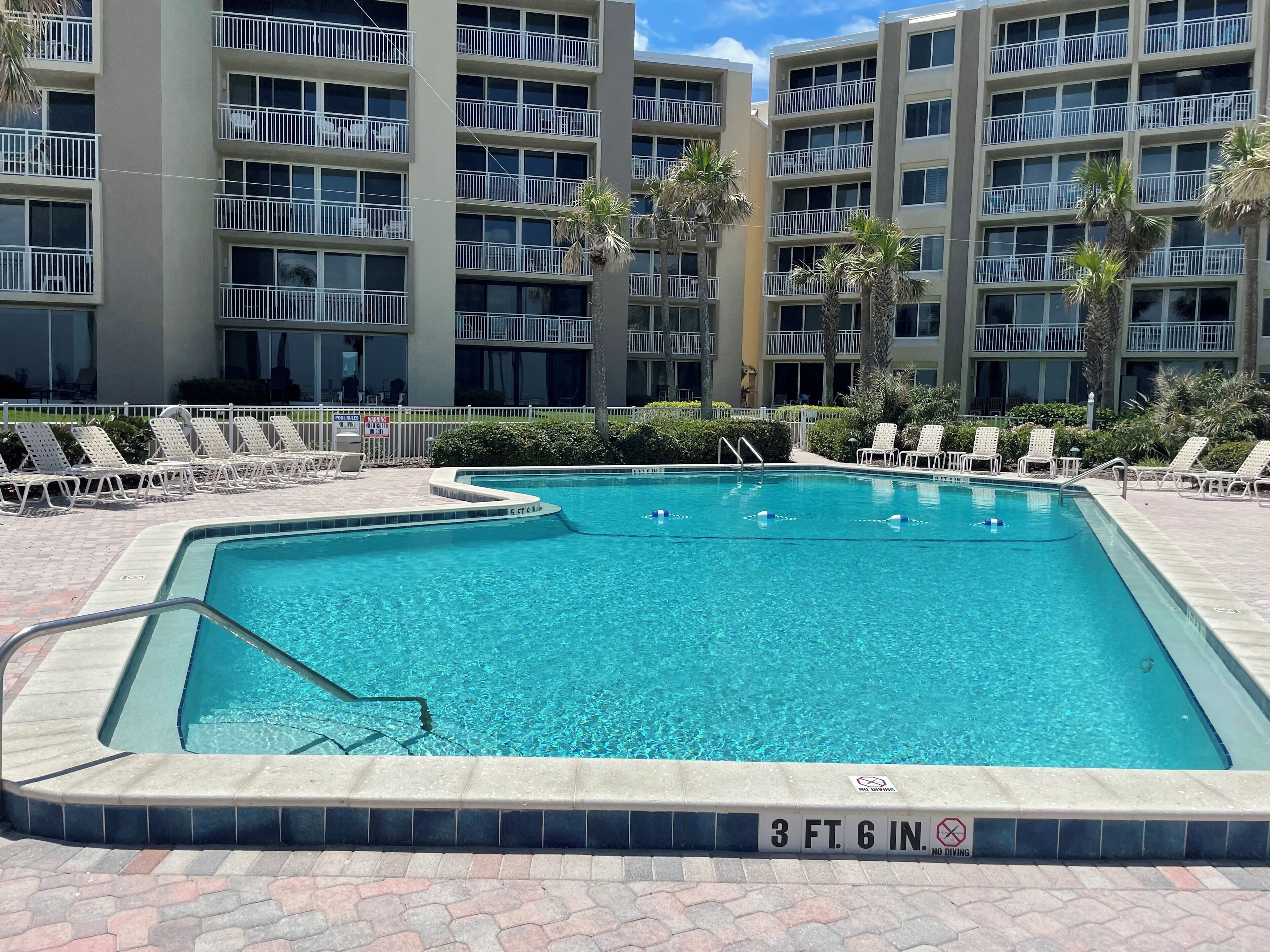 A rectangular pool with a 3 ft. 6 in. depth marker, surrounded by lounge chairs and palm trees, with a multi-story building in the background.
