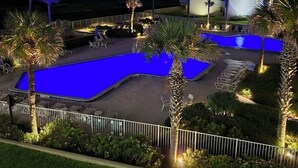 Vista nocturna de un área de piscina de hotel, con una piscina iluminada en azul, palmeras y un edificio de varios pisos al fondo.