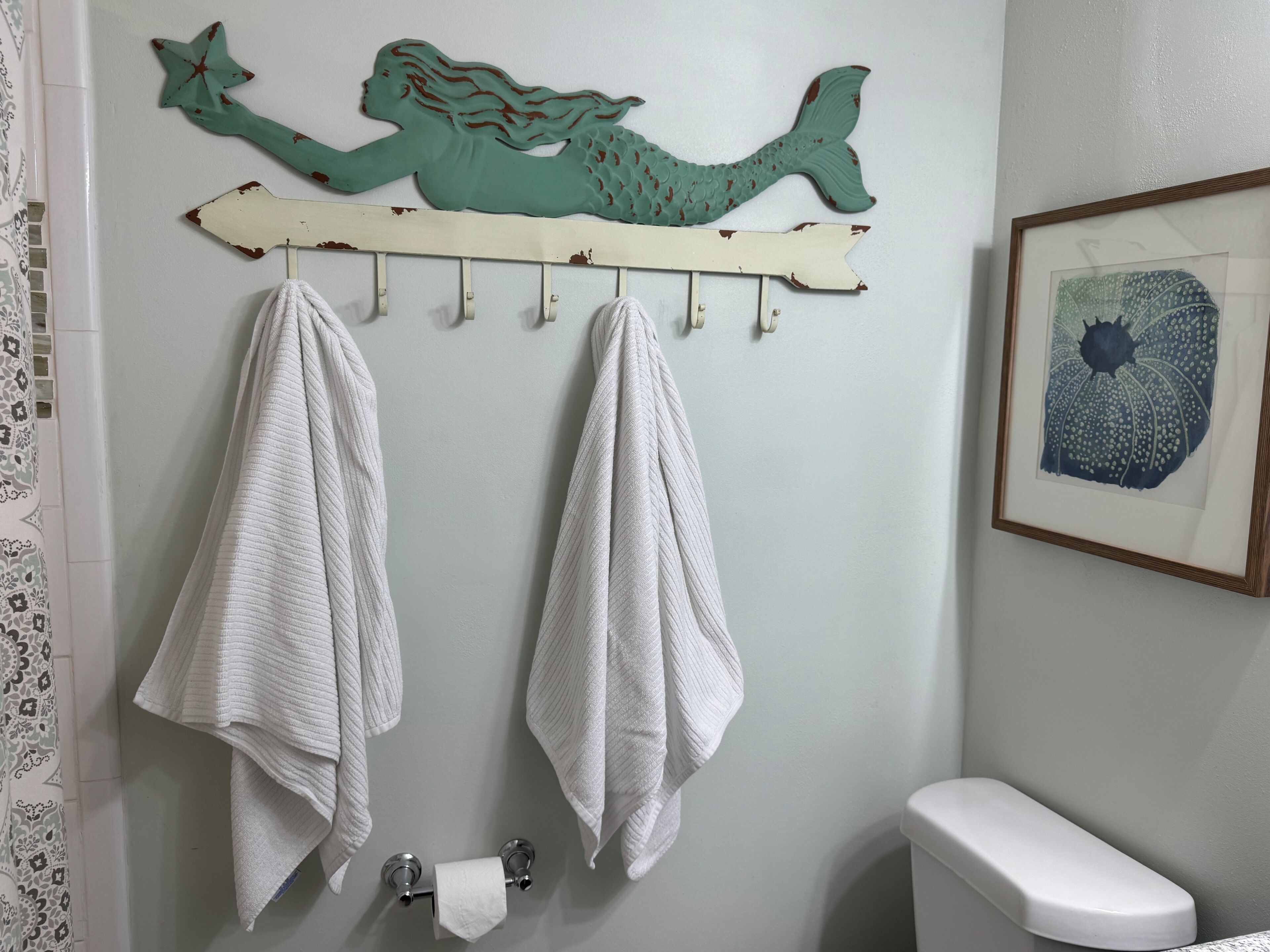 Une salle de bain décorée sur le thème des sirènes, avec deux serviettes suspendues à un porte-serviettes, des toilettes et une œuvre d’art encadrée accrochée au mur.