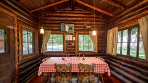 A log cabin with a dining area featuring a table with checkered tablecloths and patterned chairs, wooden walls, and large windows with curtains.