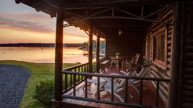A wooden cabin with a porch, outdoor seating, and a view of a lake at sunset.