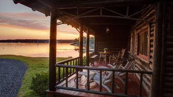 A wooden cabin with a porch, outdoor seating, and a view of a lake at sunset.