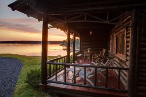 A wooden cabin with a porch, outdoor seating, and a view of a lake at sunset.