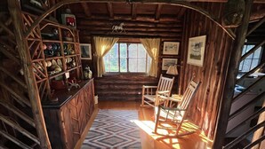 A rustic cabin interior with a wooden floor, a dining area with chairs, a bookshelf, and a window with curtains.