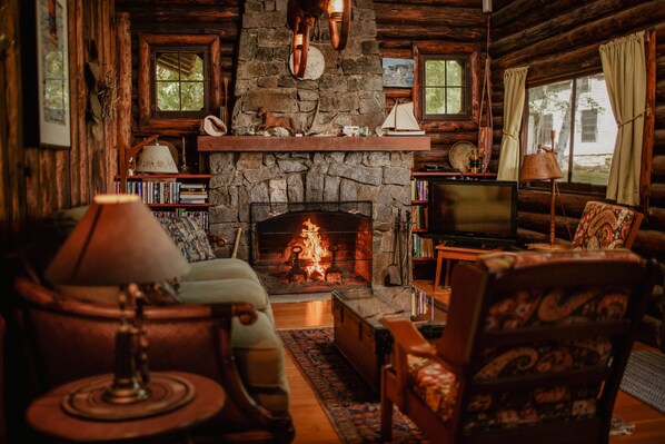 A cozy log cabin interior with a stone fireplace, wooden furniture, and a bookshelf.