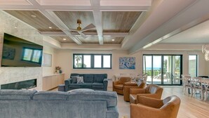 A spacious living area with a large gray sectional sofa, a dark gray couch, and several brown leather chairs. There is a flat-screen TV mounted on the wall, a fireplace, and a large window providing a view of the outdoors.