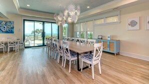 A spacious dining area with a long table and chairs, a decorative ceiling light, and large windows offering a view of the outdoors.