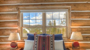 Une chambre située dans une cabane en rondins, avec une grande fenêtre, un lit recouvert d’une couette bleue et blanche, et deux tables de chevet avec des lampes.