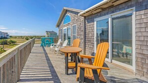 Una terraza de madera con una mesa, sillas, un ventanal y vista a la playa.