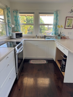 A compact kitchen with white cabinets, a microwave, an oven, and a sink. There are two windows with curtains, a potted plant, and a framed picture on the wall. - North Shore Ocean-View Garden Cottage near Paia – Maui by the Sea (Paia)
