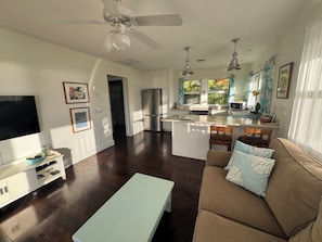 A modern living room with a flat-screen TV, a sofa with patterned cushions, a dining area with a kitchen island, and a ceiling fan. - North Shore Ocean-View Garden Cottage near Paia – Maui by the Sea (Paia)