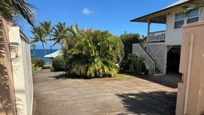 A residential area with a paved driveway, palm trees, and a white building with a balcony. - North Shore Ocean-View Garden Cottage near Paia – Maui by the Sea (Paia)