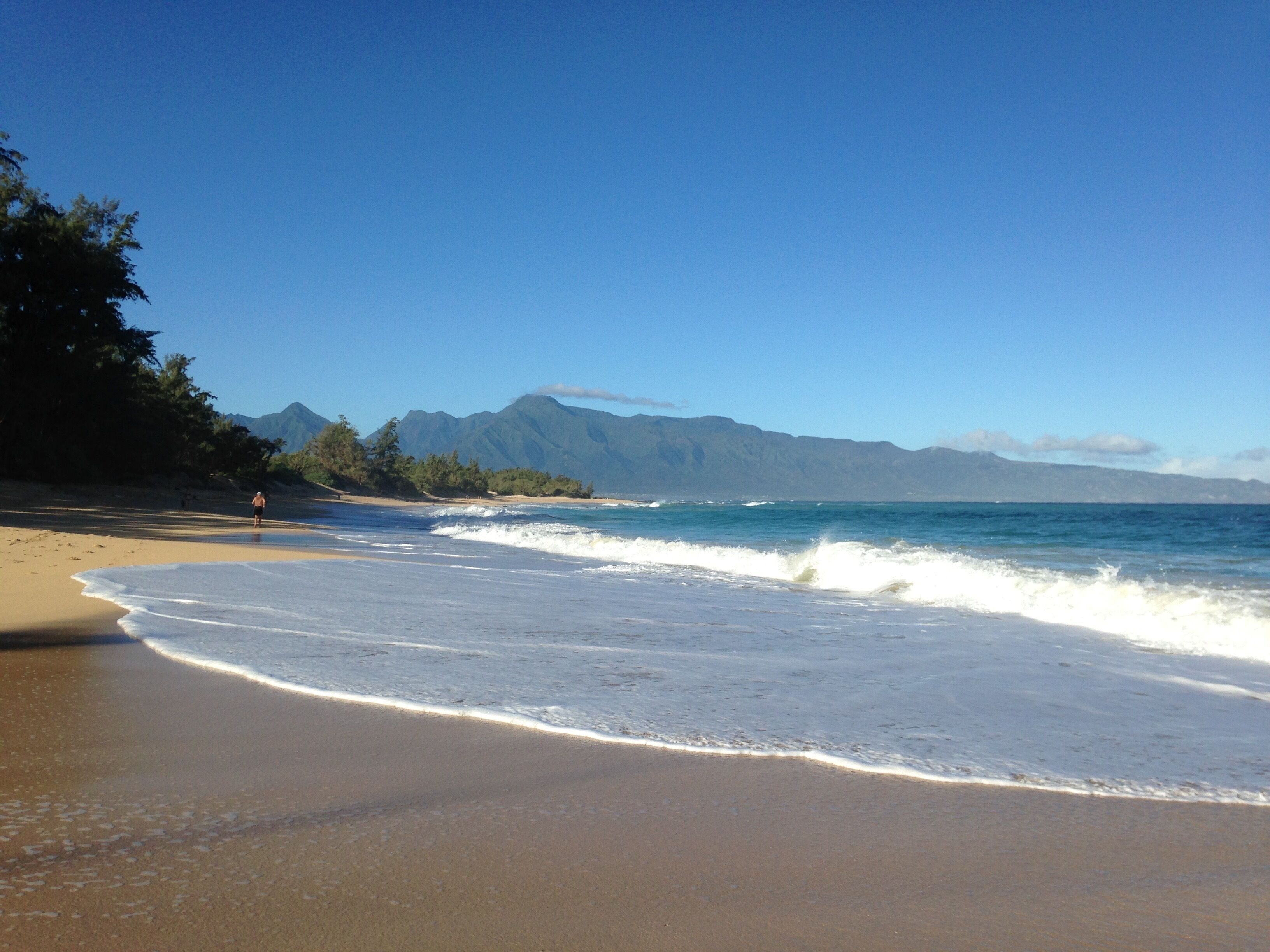 Platja amb cel blau, onades i muntanyes al fons.