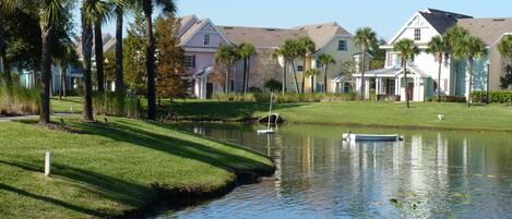 A serene lakeside view with a small boat, palm trees, and a row of houses with a pond in the foreground.