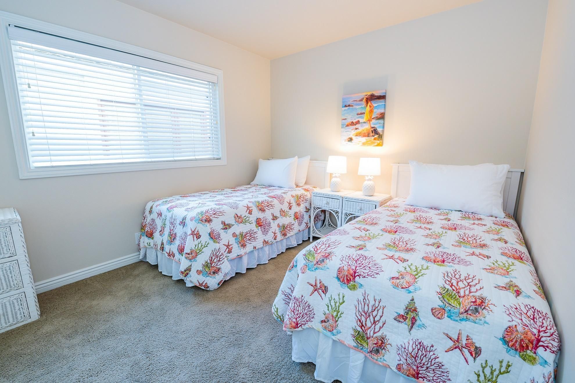 Ein Schlafzimmer mit zwei Betten, jeweils mit einem floralen Bettüberwurf, einem Fenster mit Jalousien, einem Wandbild und einem weißen Rattan-Nachttisch mit Lampen.