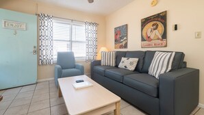 A room with a light blue door marked 'ENJOY 3', a grey sofa with striped pillows, a small table with a laptop, and a window with patterned curtains.