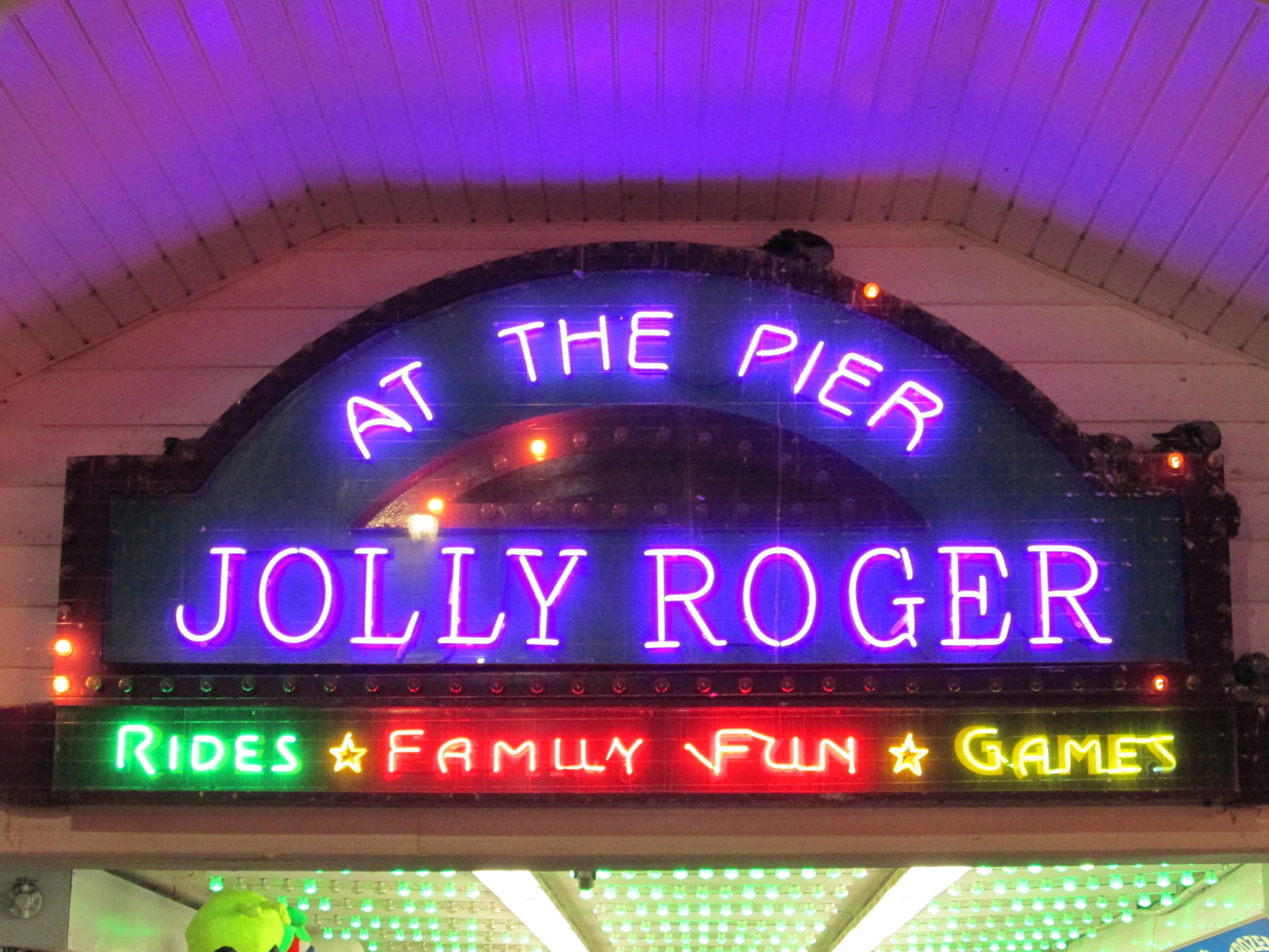 A neon sign with the text "AT THE PIER JOLLY ROGER" and below it, "RIDES FAMILY FUN GAMES".