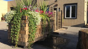 Een houten plantenbak met verschillende bloemen en planten, voor een gebouw met een bakstenen gevel en een raam.