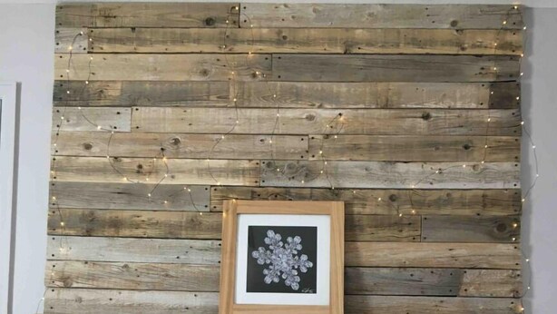 Een bed met een gewatteerde witte dekbedovertrek, een gewatteerd grijs kussen, een blauw kussen, en een kussen met een rood-zwarte geruit patroon. Boven het bed een houten hoofdbord met een ingelijste afbeelding van een sneeuwvlok.
