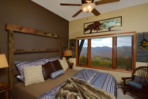 Une chambre à coucher avec un grand lit, un ventilateur de plafond, une fenêtre donnant sur les montagnes et une tête de lit en bois.
