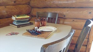 Interior de uma cabana de madeira com uma mesa redonda, cadeiras, uma pilha de livros, um prato e um jarro.